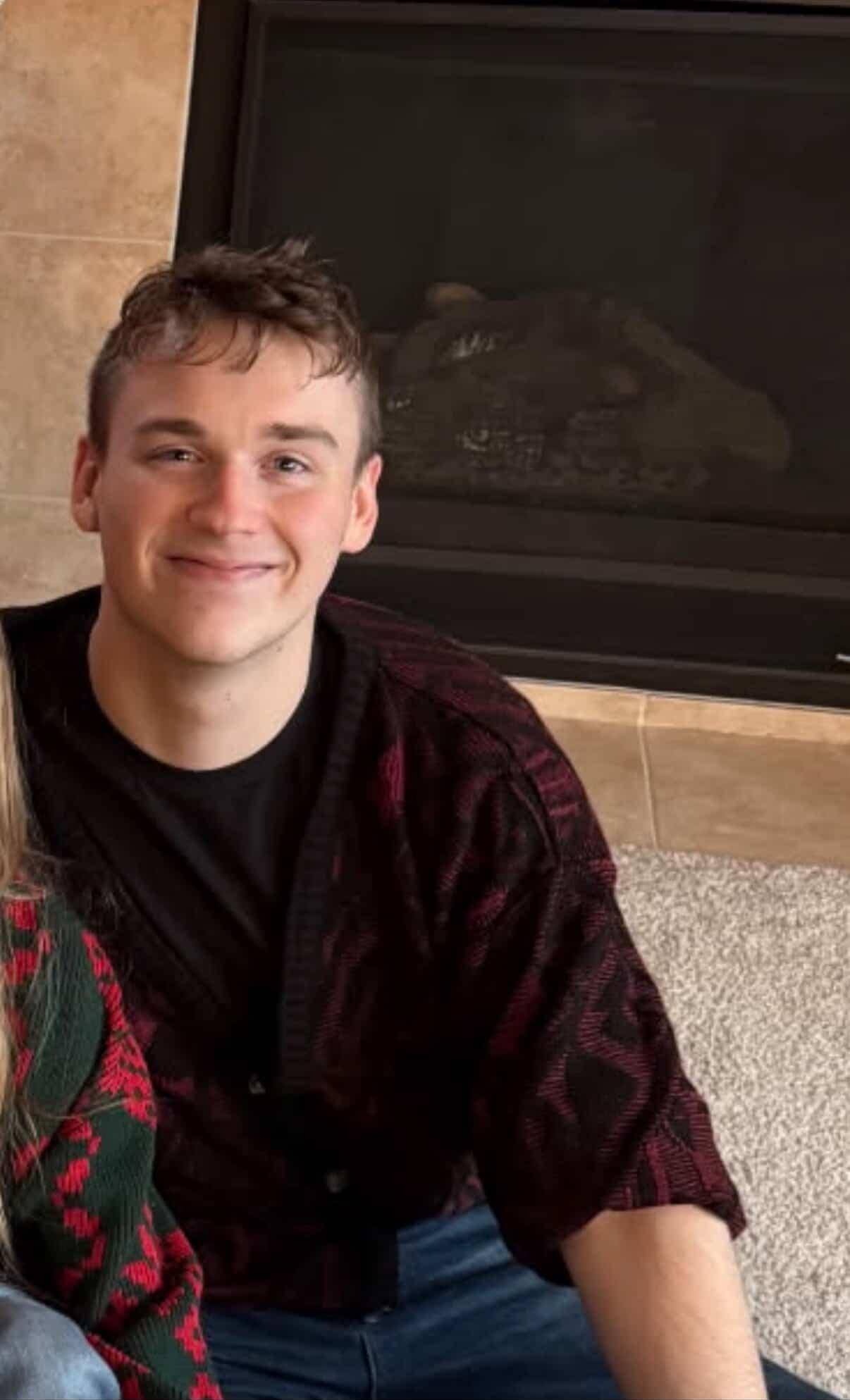 Young man with red and brown cardigan smiling at the camera with a fireplace in the background